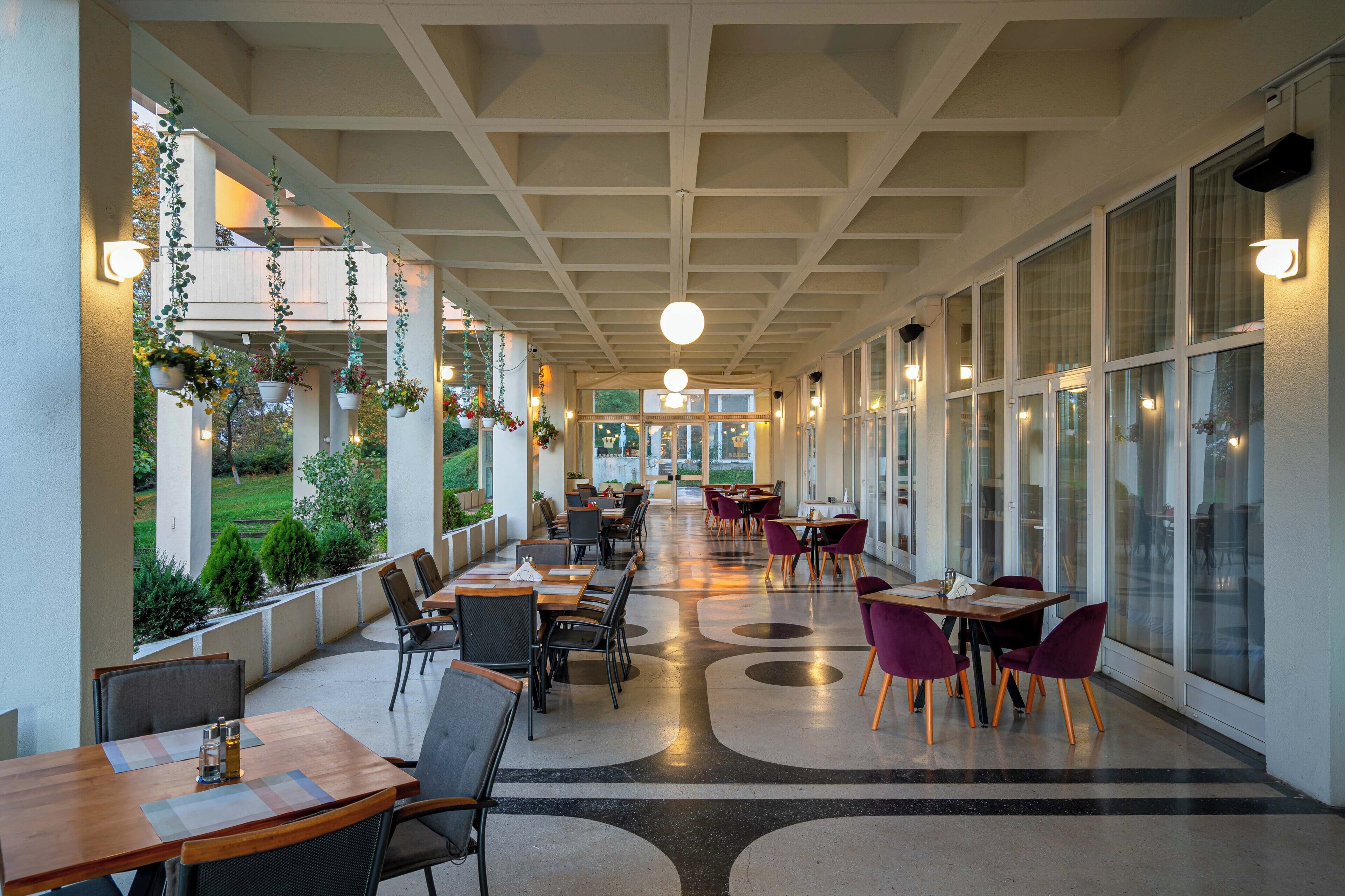 A covered outdoor dining area with tables and chairs, hanging plants, and large windows.