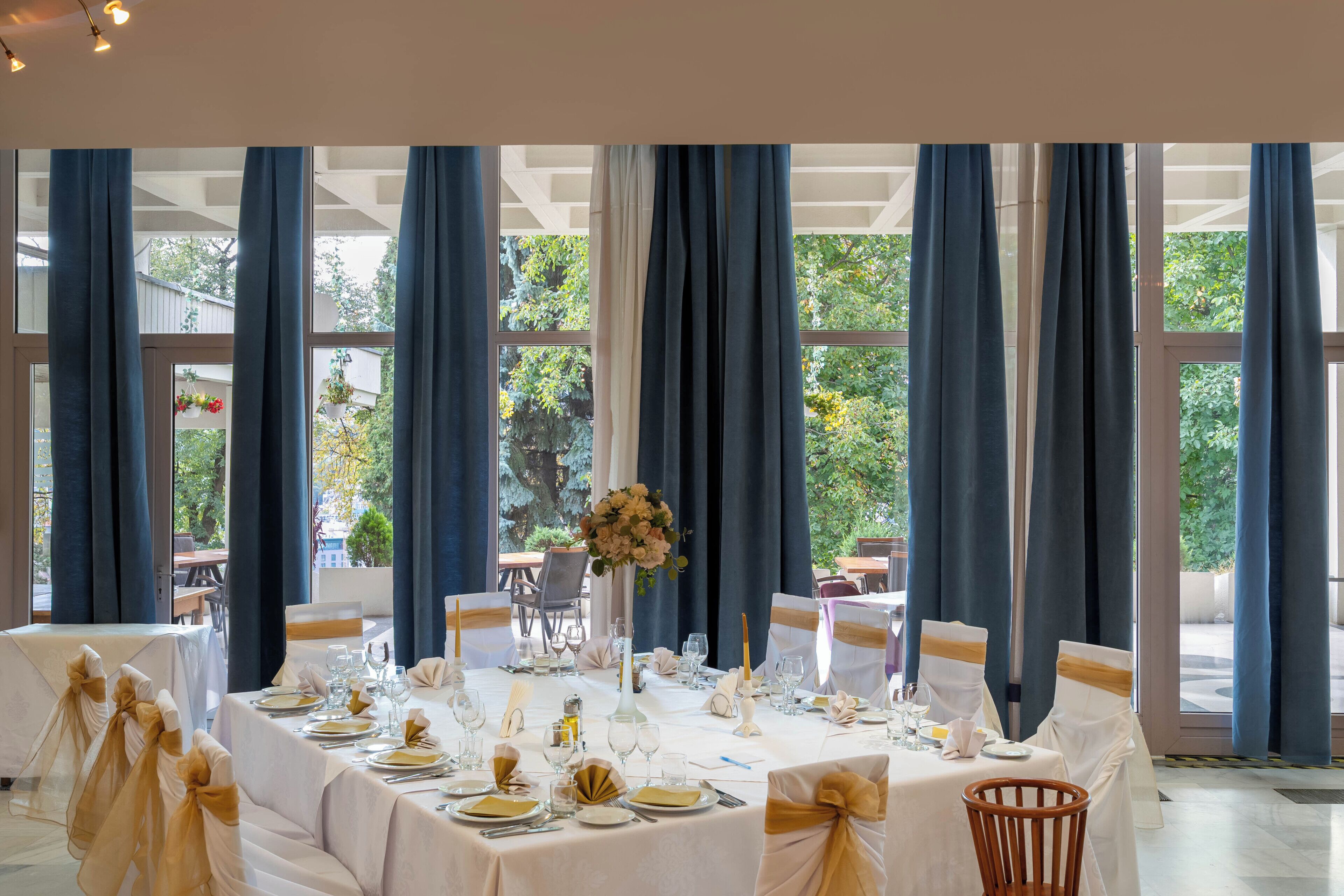 Comedor formal con mesas dispuestas para una comida, ventanas amplias con cortinas y vistas a follaje exterior.