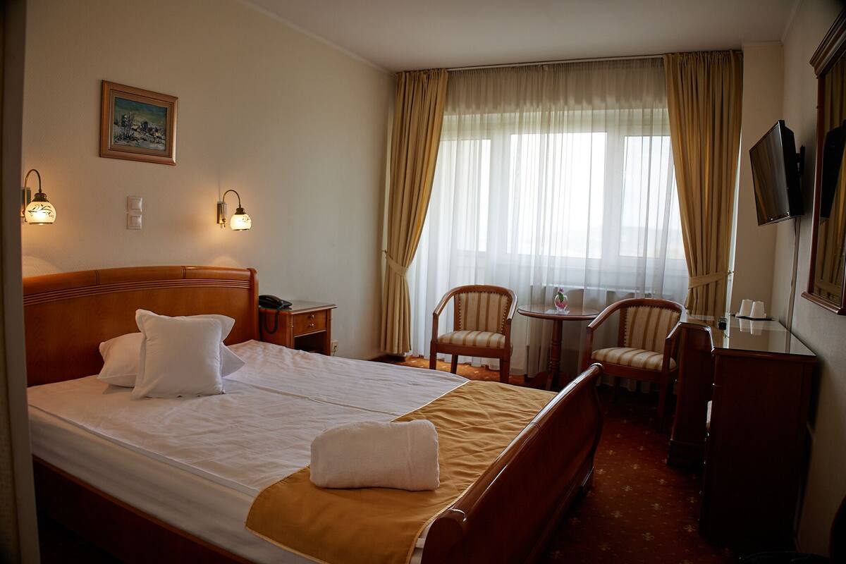 A hotel room with a large bed, two chairs, a small table, a TV, and a painting on the wall.