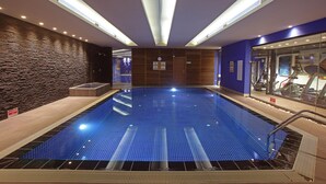 A swimming pool with a stone wall and a blue tiled floor, surrounded by a wooden deck and a glass wall with exercise equipment. - Wes Hotel Izmit (Izmit)