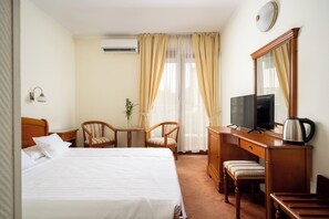 A hotel room with a bed, a desk with a television, a chair, a small table, and a window with curtains.