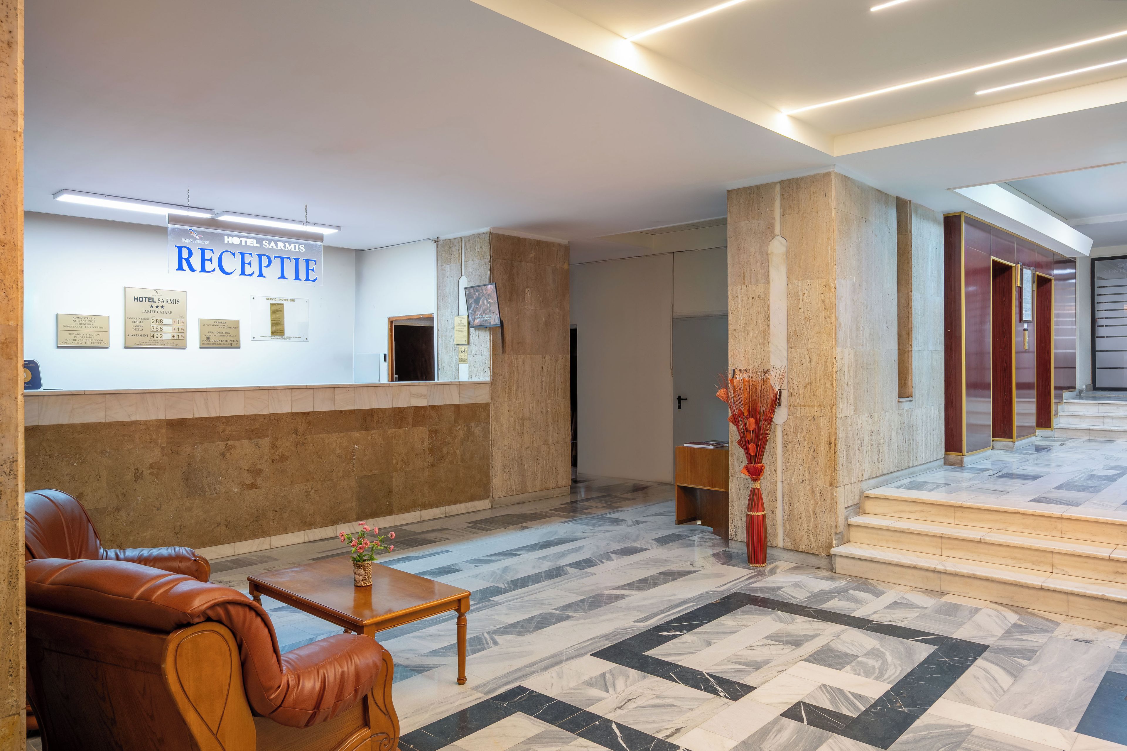 A hotel reception area with a reception desk, a leather sofa, a coffee table, and a staircase leading to other floors.