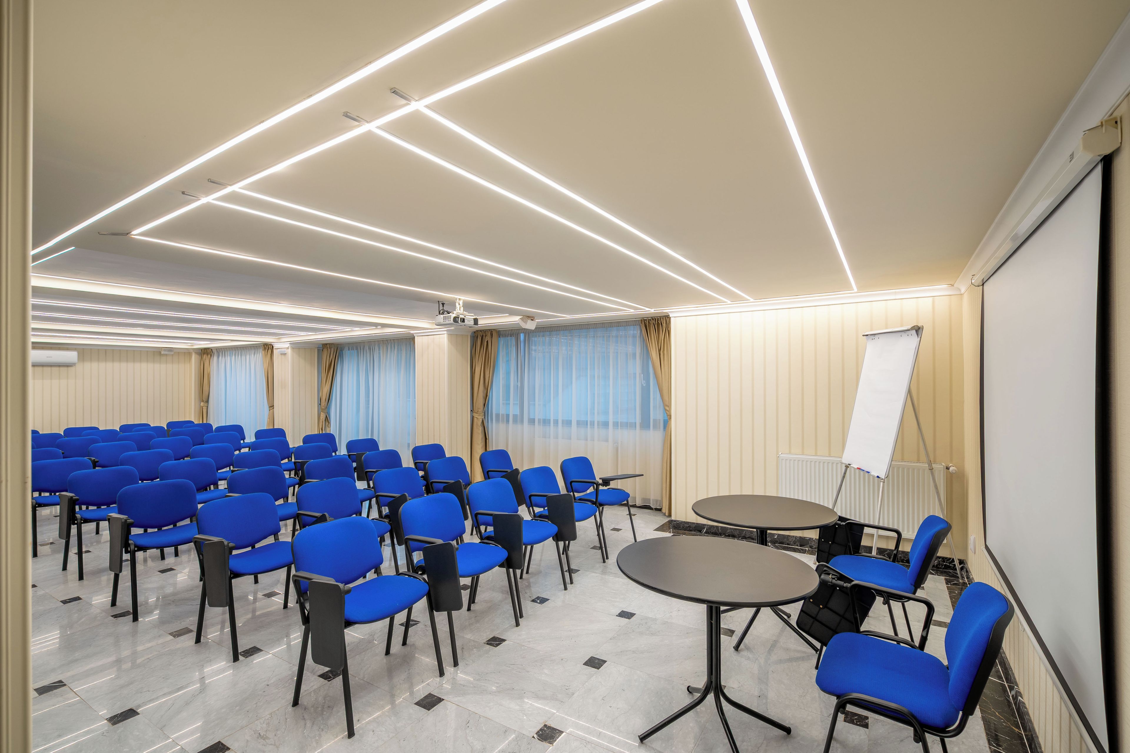 Un salón de conferencias con filas de sillas azules, una pantalla de proyector y una pizarra.