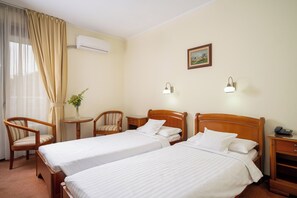 A hotel room with two single beds, a wooden chair, a small table with a vase, and a painting on the wall.