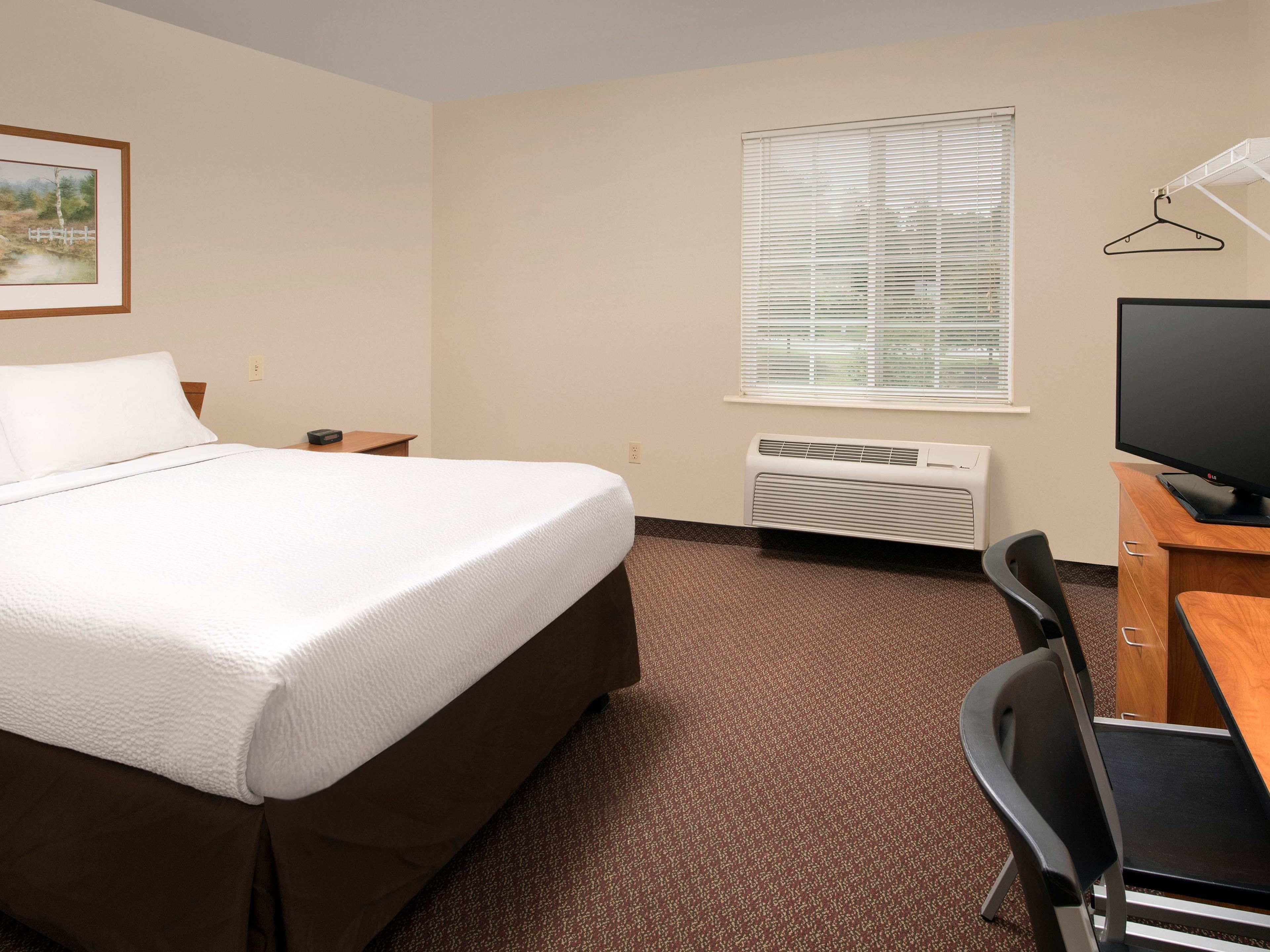 Una habitación de hotel con una cama, un escritorio con silla, un televisor, una ventana con persianas y un equipo de aire acondicionado.