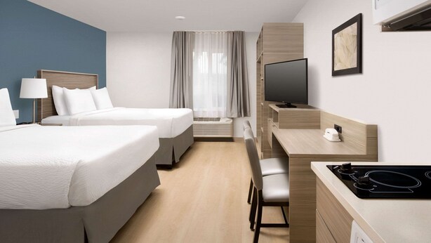 A hotel room with two beds, a desk with a TV, a chair, and an air conditioning unit. - WoodSpring Suites Charlotte Gastonia (Gastonia)