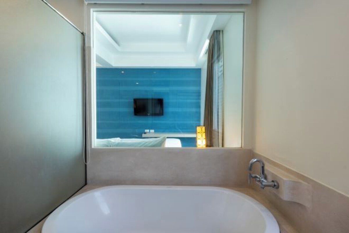 Un baño moderno con bañera, ducha y un espejo grande que refleja un cuarto con cama y televisor.