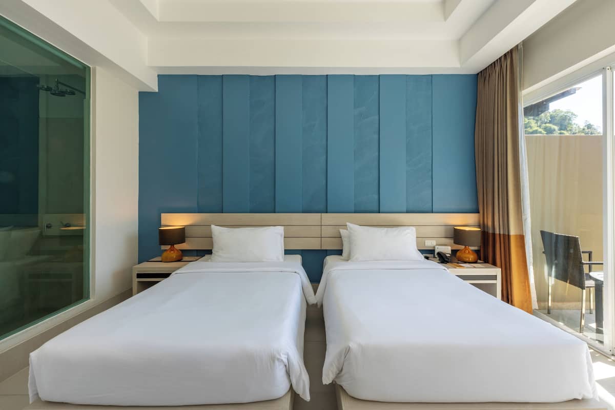 A hotel room with two single beds, a large window with curtains, a nightstand with a lamp, and a glass shower enclosure.