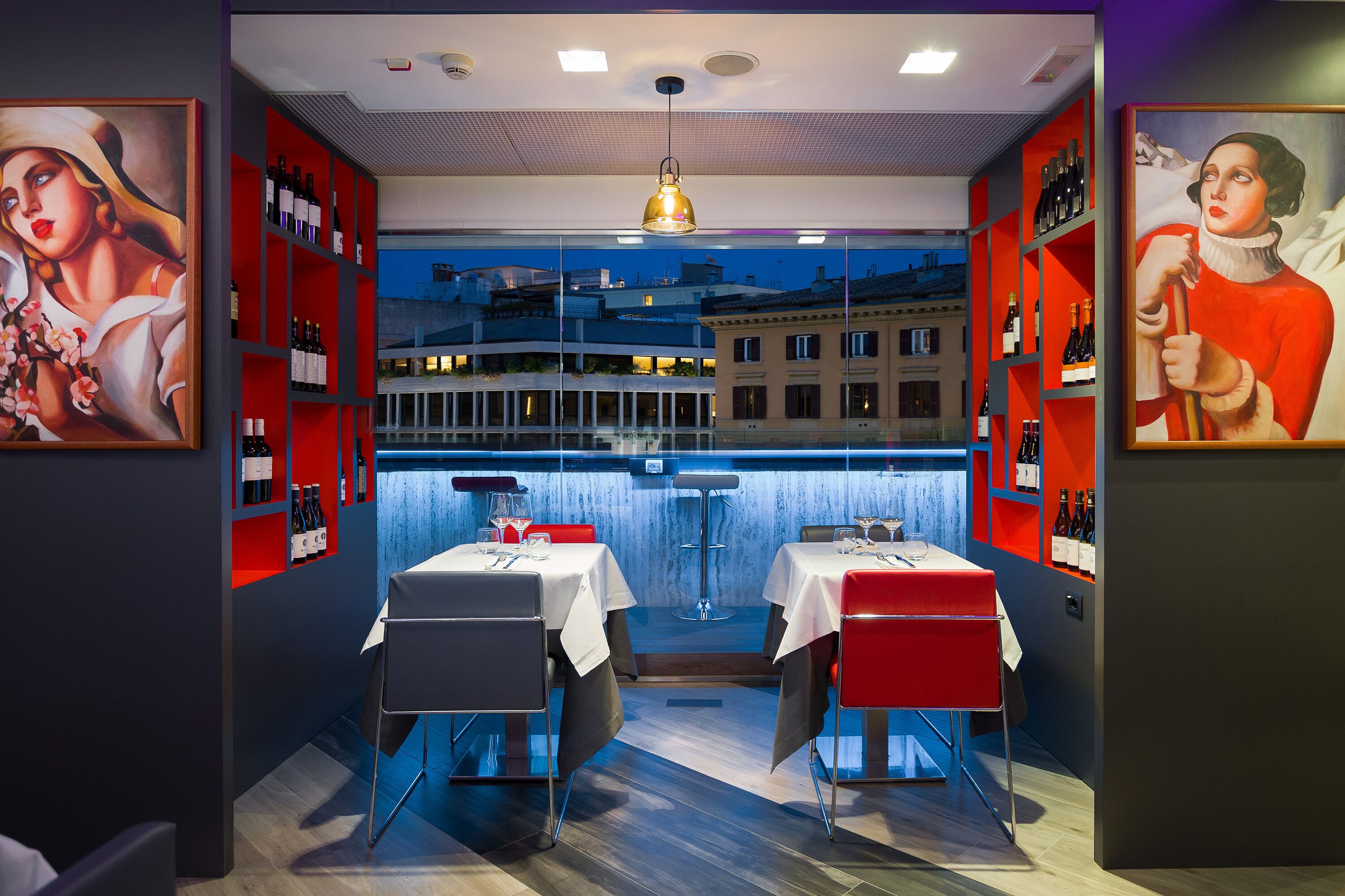 Un restaurante con dos mesas, sillas rojas y grises, y botellas de vino en estanterías.