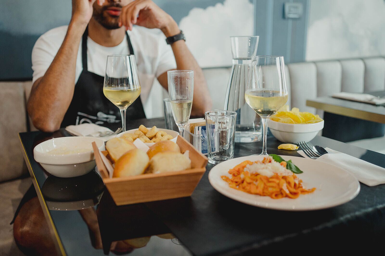 A dining table with a plate of pasta, a bowl of bread, and glasses of white wine.