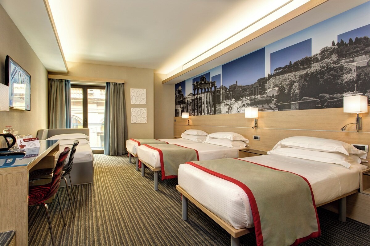 Habitación de hotel con tres camas, un escritorio, sillas, un televisor y un mural grande en la pared.