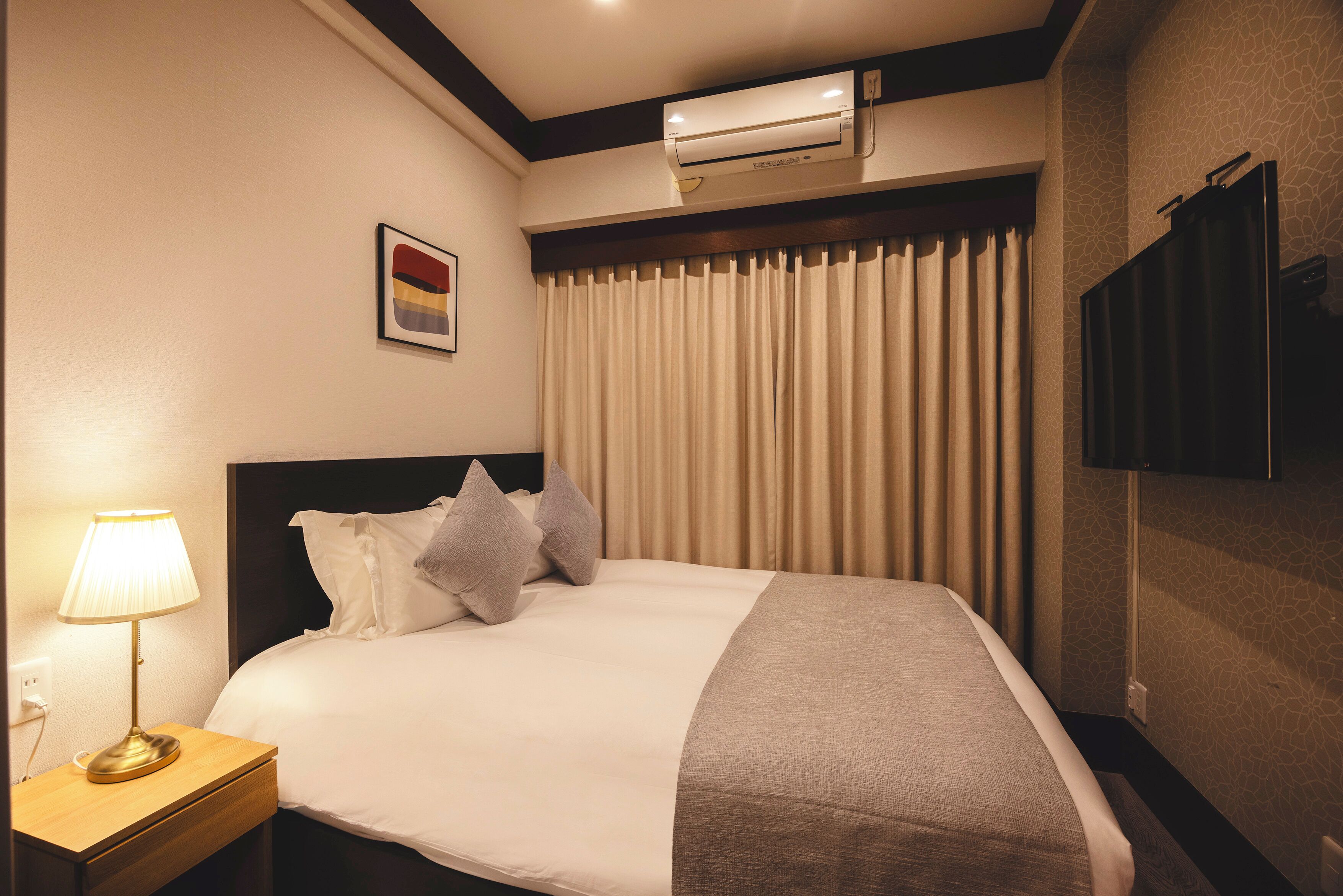 A hotel room with a bed, a nightstand with a lamp, a wall-mounted air conditioner, and a television on the wall.