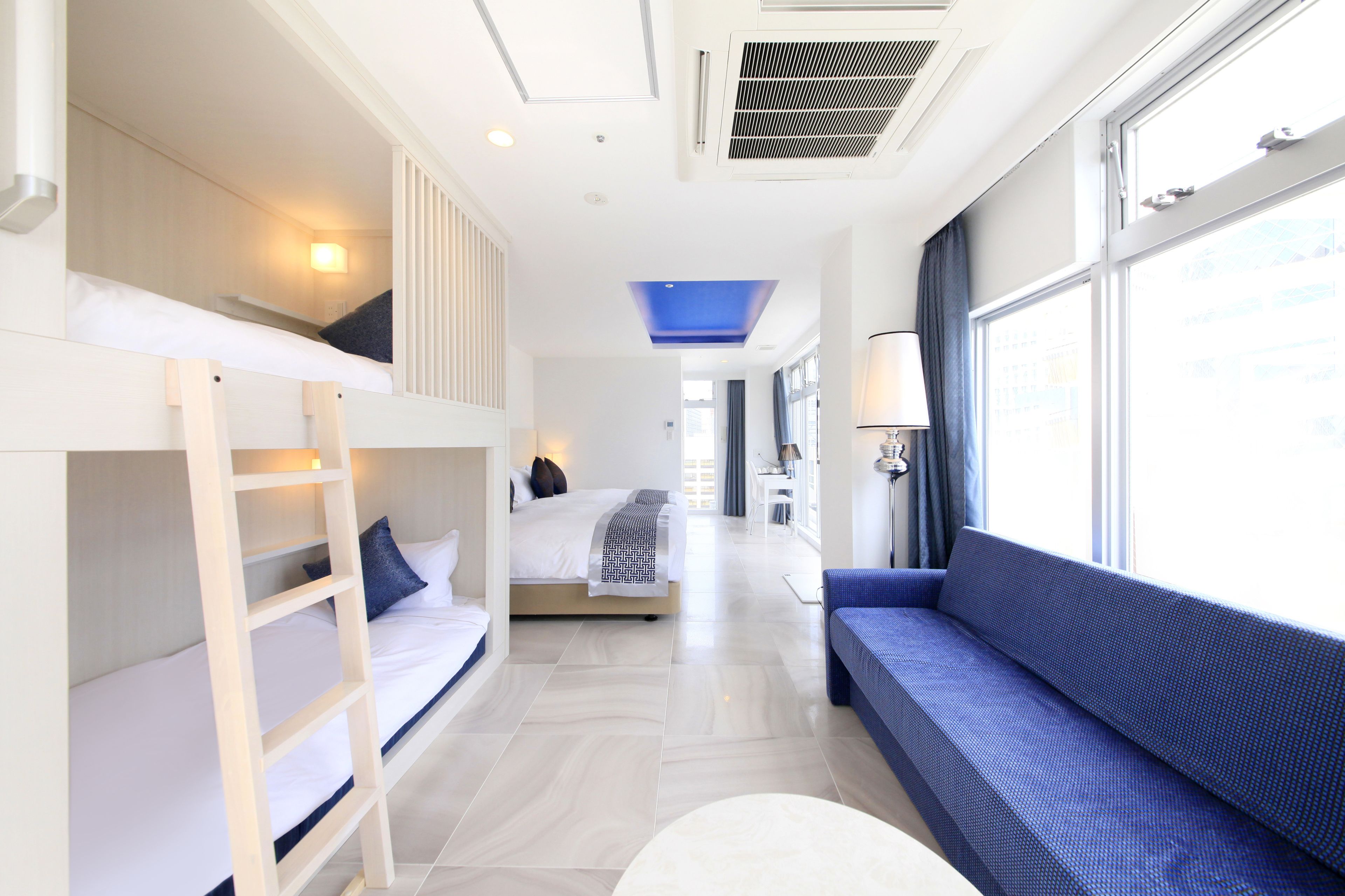 A modern hotel room with a bunk bed, a sofa, a round table, and a large window providing a view of the city.