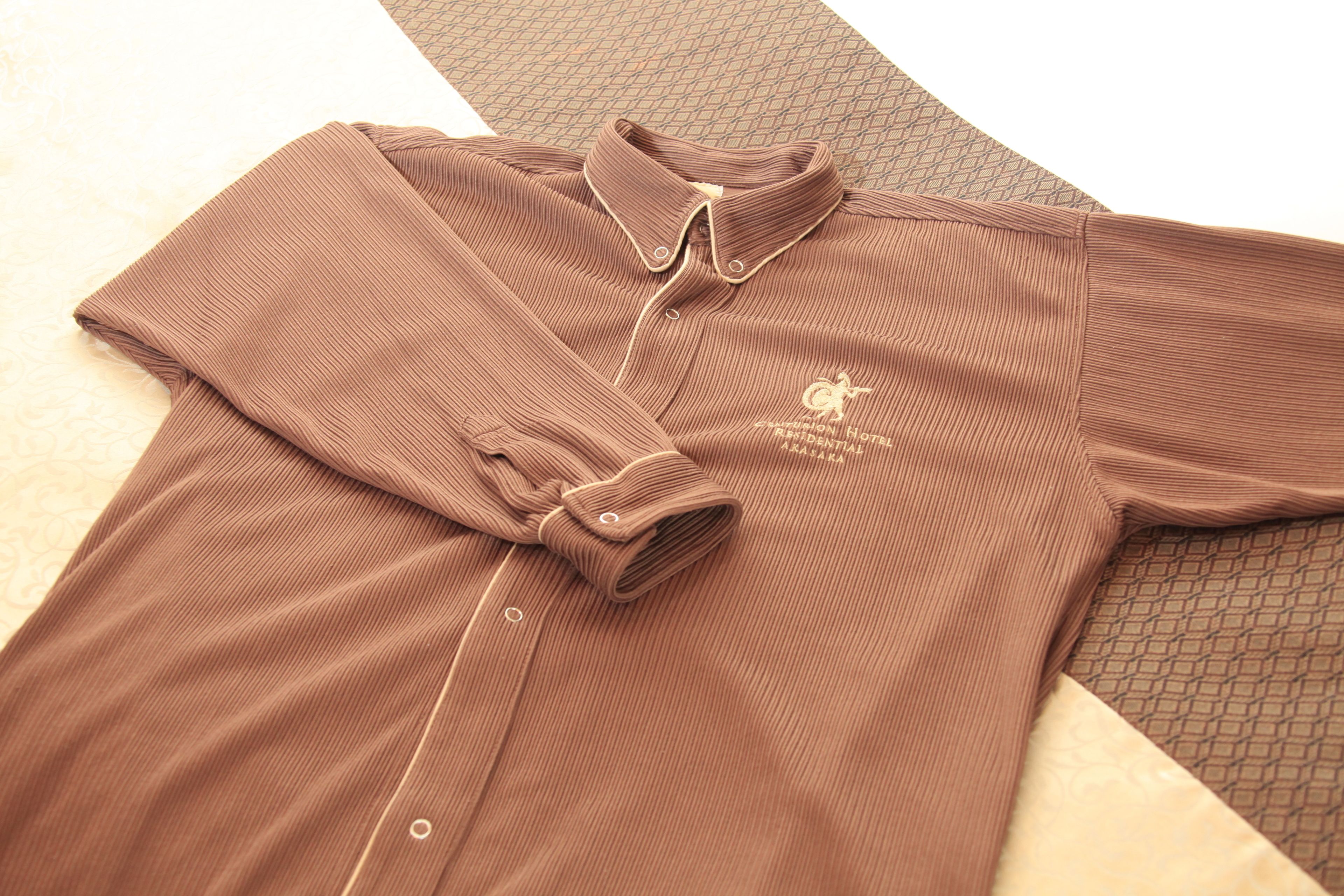 Ein braunes Poloshirt mit einem Logo auf der linken Brust, ausgebreitet auf einer gemusterten Fläche.