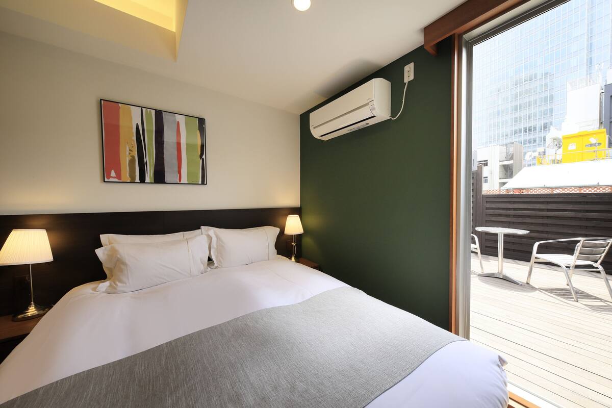 Une chambre d’hôtel avec un grand lit, deux lampes de chevet, un climatiseur fixé au mur, un tableau abstrait aux couleurs vives, et une vue sur la ville par une grande fenêtre.