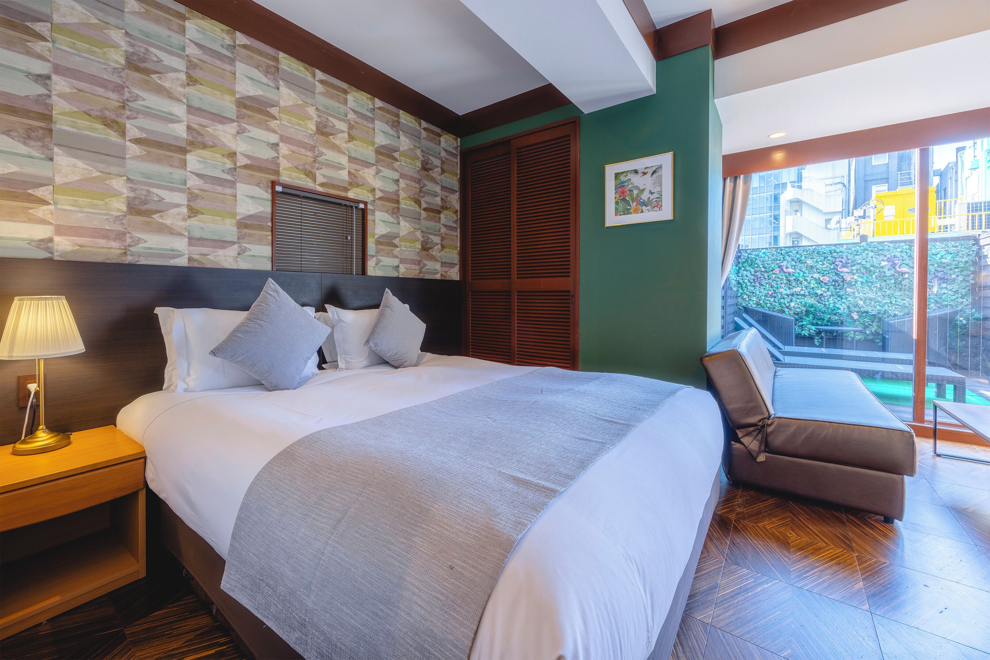 A hotel room with a large bed, a nightstand, a window with a view of buildings, and a balcony with plants.