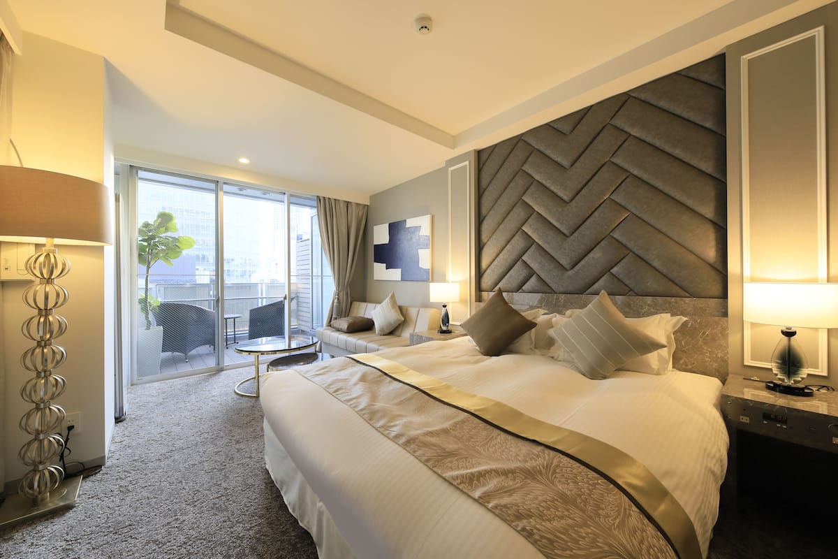 Une chambre d’hôtel moderne dotée d’un grand lit, d’une tête de lit à motifs en chevrons, d’un balcon avec mobilier d’extérieur et offrant une vue sur le panorama urbain.