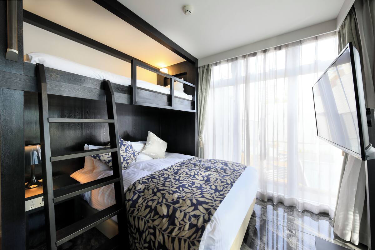 Une chambre d’hôtel avec un lit superposé, un téléviseur à écran plat et une grande fenêtre avec des rideaux légers.