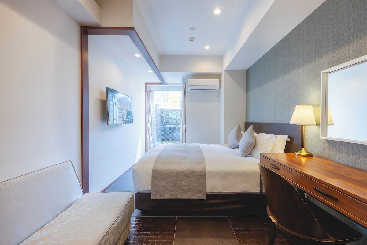 Une chambre d’hôtel avec un lit, un bureau en bois, une lampe et une vue sur l’extérieur, visible à travers une porte.