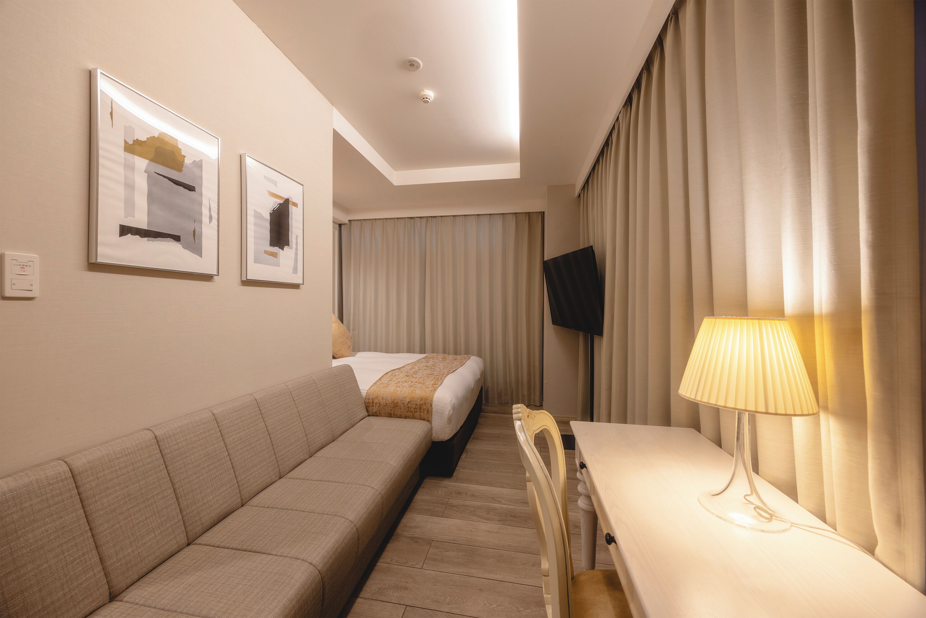 A hotel room with a bed, a sofa, a desk, a lamp, and a TV on the wall.