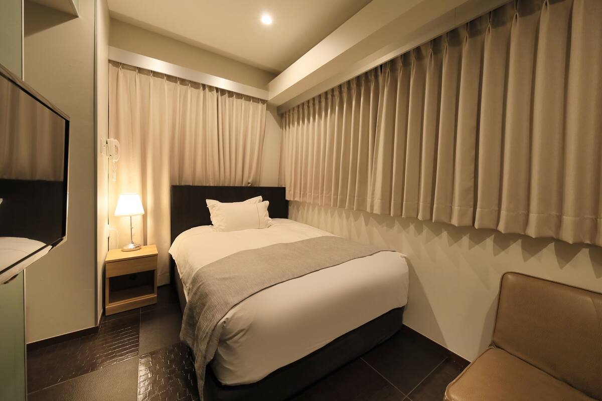 Une chambre d’hôtel avec un lit, une table de chevet avec une lampe, une chaise et une grande fenêtre avec des rideaux.