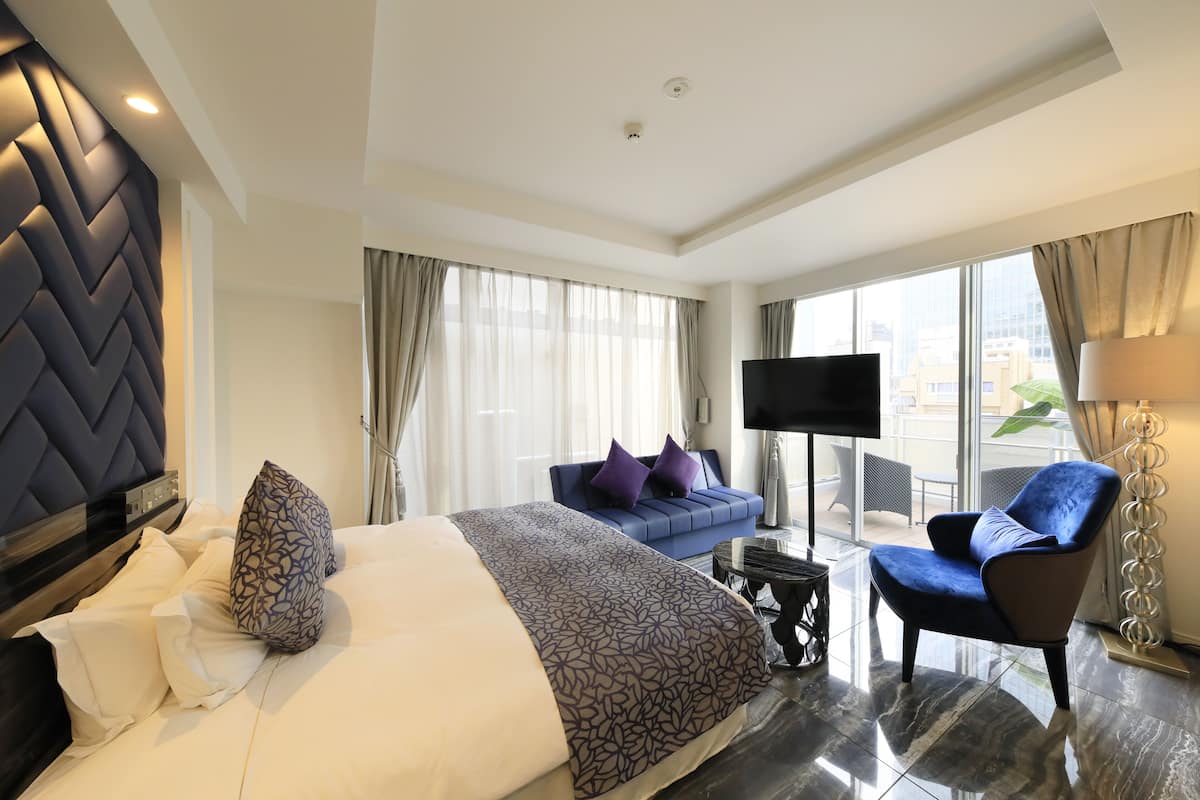 Une chambre d’hôtel moderne avec un grand lit, un canapé bleu, un téléviseur à écran plat et une vue sur la ville grâce à de grandes fenêtres.
