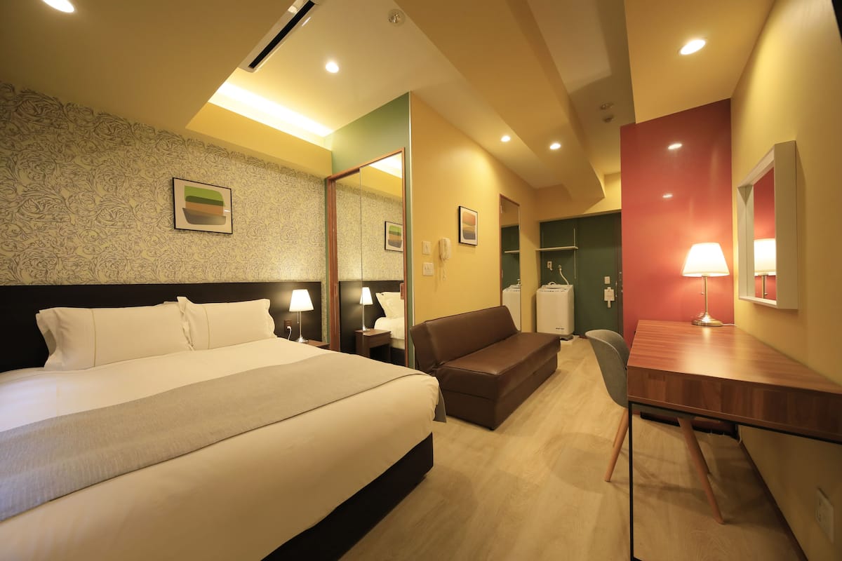 Une chambre d’hôtel avec un grand lit, un bureau, une chaise, un canapé, un miroir et une salle de bain visible à travers une porte ouverte.