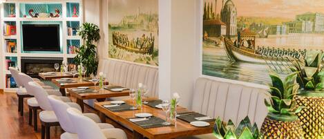 A restaurant with long wooden tables, white cushioned chairs, and a large mural of a river scene.