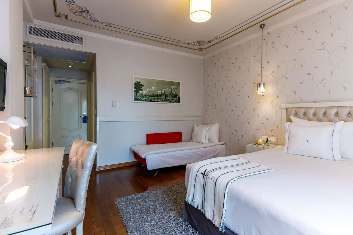 Une chambre d’hôtel avec un grand lit, un lit plus petit, un bureau avec une chaise, un mur d’accent rouge et un tableau encadré accroché au mur.