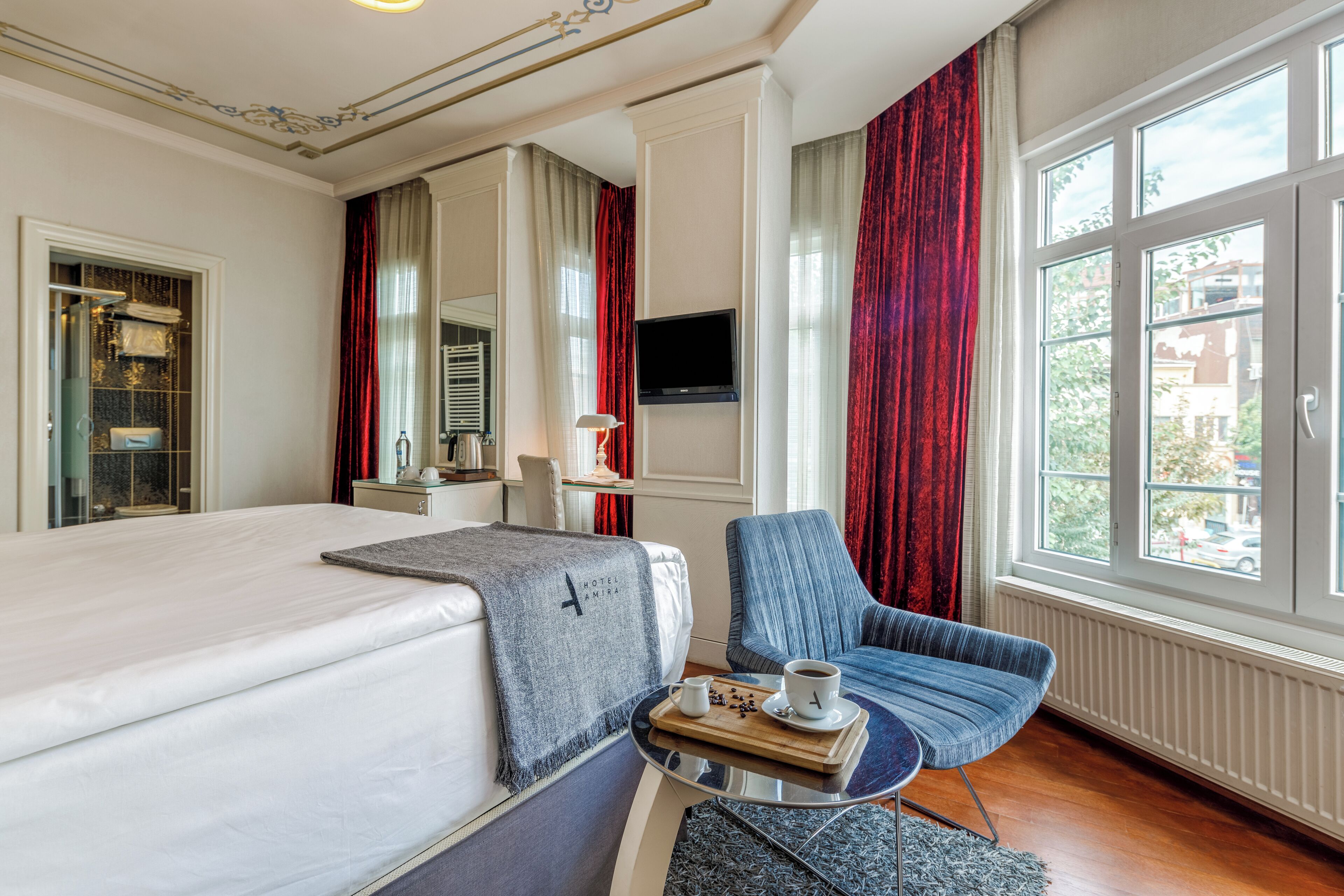 Ein Hotelzimmer mit einem Bett, einer Sitzecke mit Tisch, einem Fernseher und großen Fenstern mit Vorhängen.