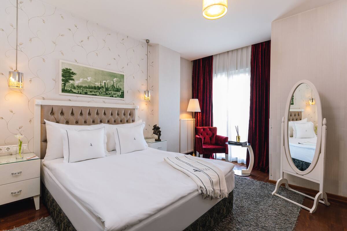 Une chambre d’hôtel avec un grand lit, un fauteuil rouge, un miroir et un tableau accroché au mur.