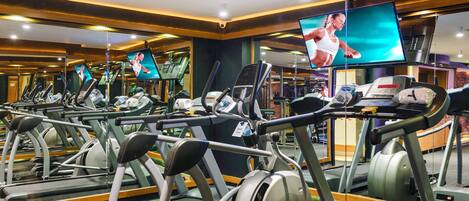 A gym with multiple treadmills, exercise bikes, and a large TV screen displaying a fitness video.