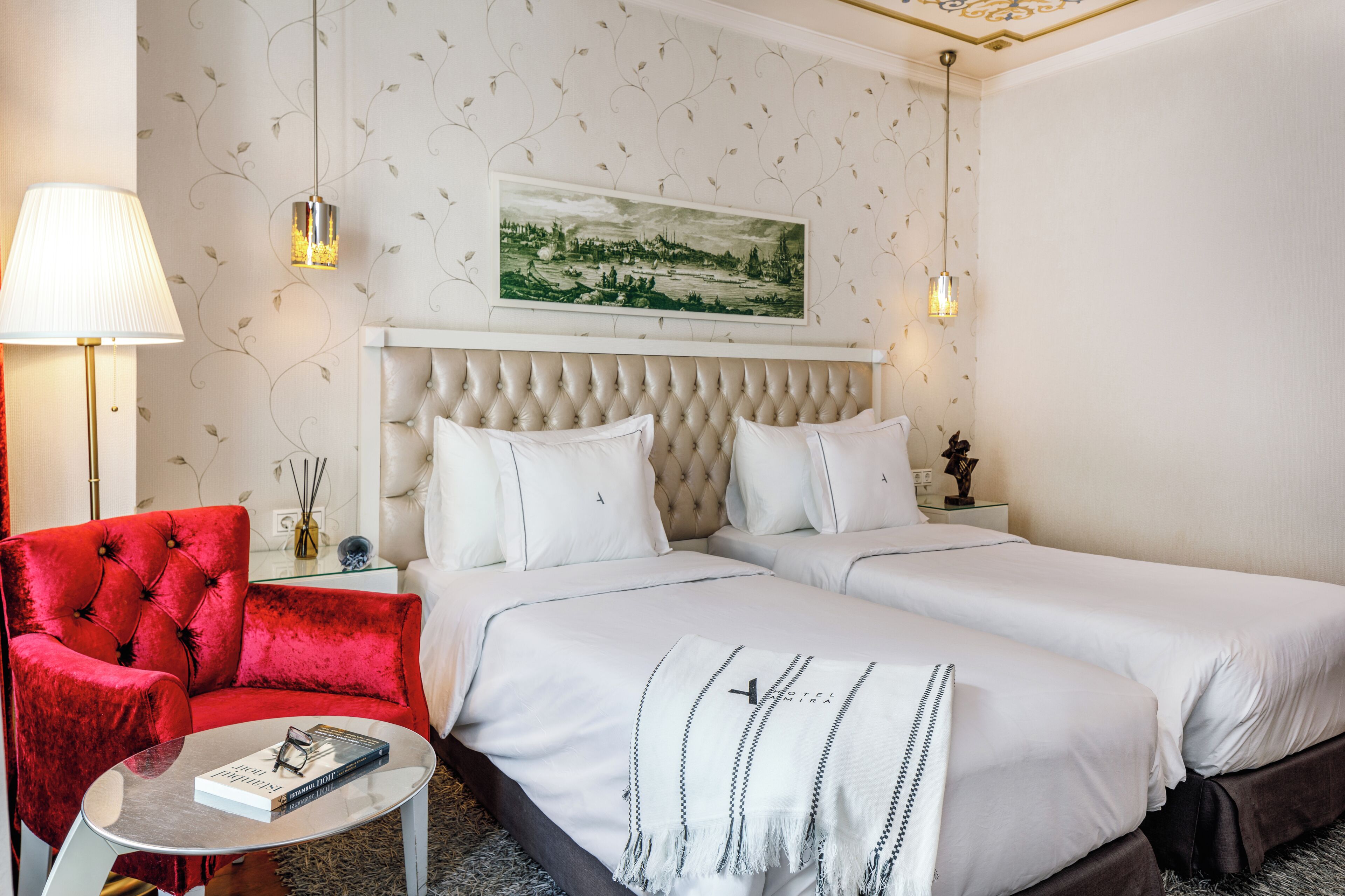 A hotel room with a large bed, a red armchair, a small table with a lamp, and a framed picture on the wall.