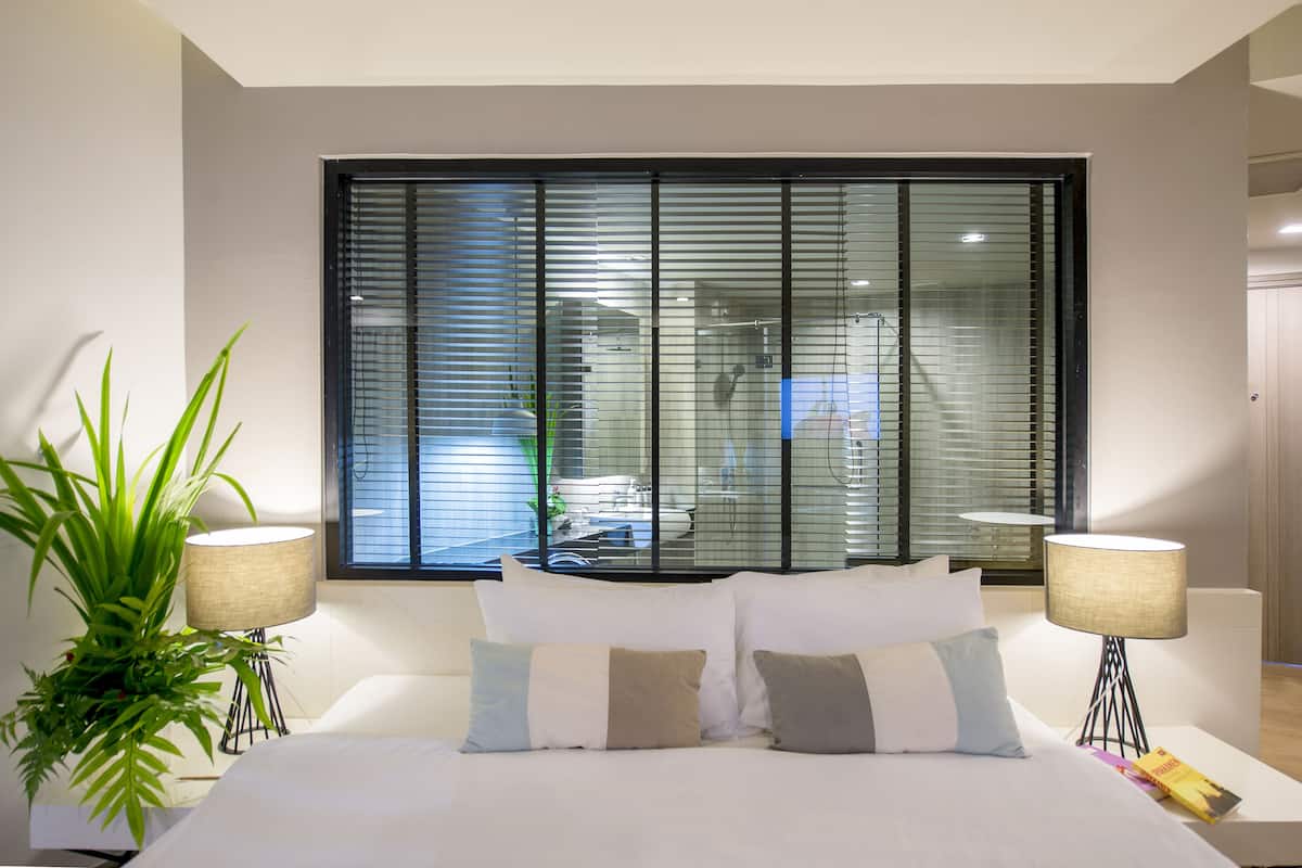 Une chambre d’hôtel moderne avec un grand lit, deux lampes de chevet, une plante en pot et une grande fenêtre avec des stores.