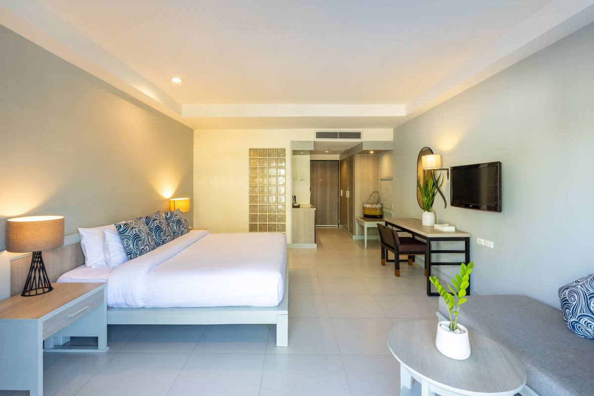 Une chambre d’hôtel moderne équipée d’un lit, d’une table de chevet, d’une lampe, d’une télévision, d’un coin salon et d’une petite table agrémentée d’une plante.