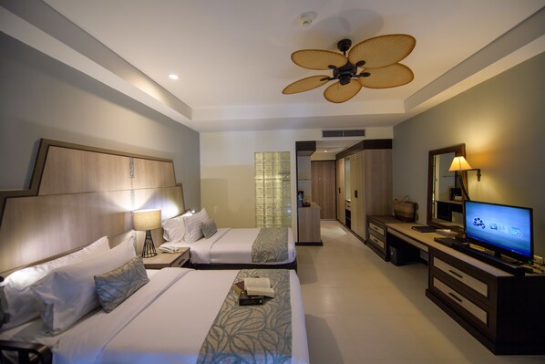 Habitación de hotel con dos camas, televisor, escritorio y ventilador de techo.