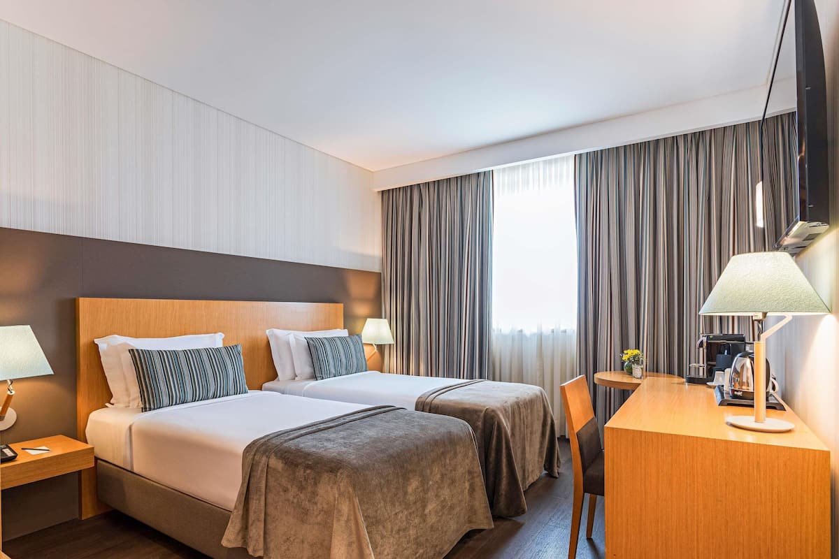 A hotel room with two beds, a desk with a coffee maker, and a view of the city through large windows.