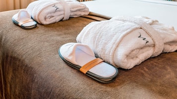 Una cama bien hecha con un edredón blanco y una almohada a rayas, un par de pantuflas y una toalla enrollada sobre una colcha marrón texturizada.