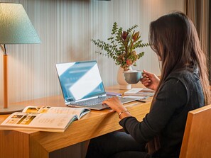 A person sitting at a wooden desk with a laptop, a book, and a cup. - Mercure Lisboa Almada (Almada)