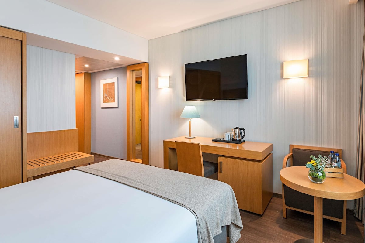 A hotel room with a bed, a desk, a chair, a television, and a small table with a lamp and a plant.