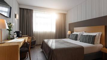 Habitación de hotel con una cama grande, un escritorio con silla, un televisor montado en la pared y una ventana con cortinas.