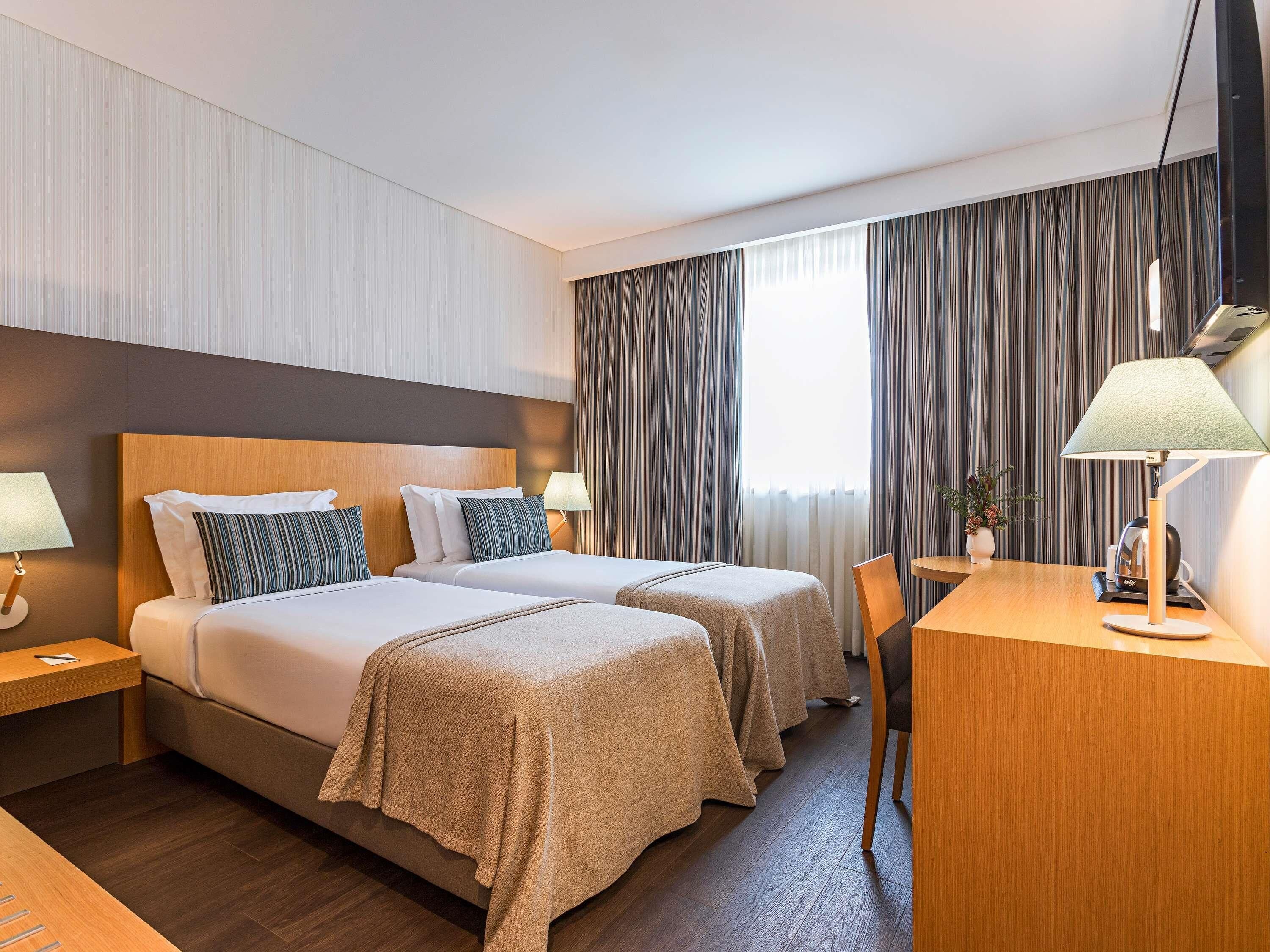 A hotel room with two beds, a desk, a chair, a lamp, and a large window with curtains.
