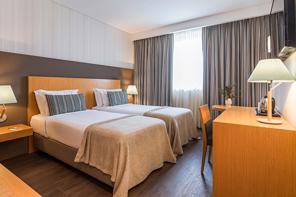 Habitación de hotel con dos camas, un escritorio, una silla, una lámpara y un ventanal grande con cortinas.