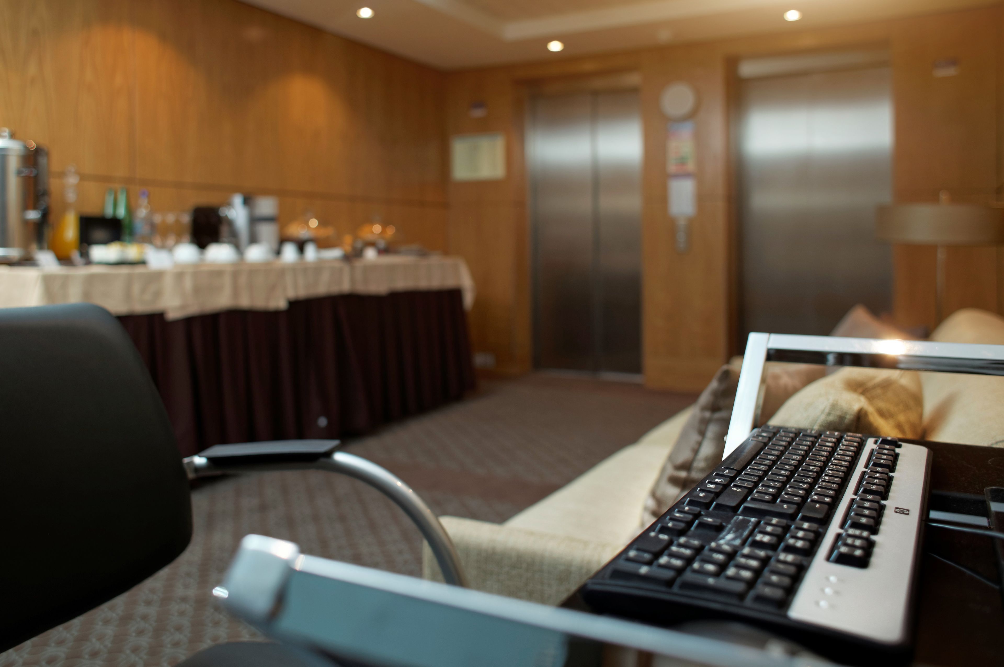 Une chambre d'hôtel équipée d'un bureau, d'un ordinateur portable, d'une chaise, d'une table recouverte d'une nappe, d'une machine à café, et avec un ascenseur visible en arrière-plan.