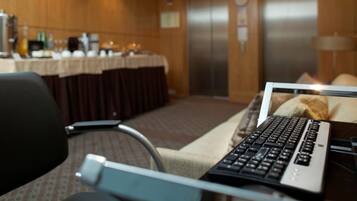 A hotel room with a desk, a laptop, a chair, a table with a tablecloth, a coffee machine, and an elevator in the background.