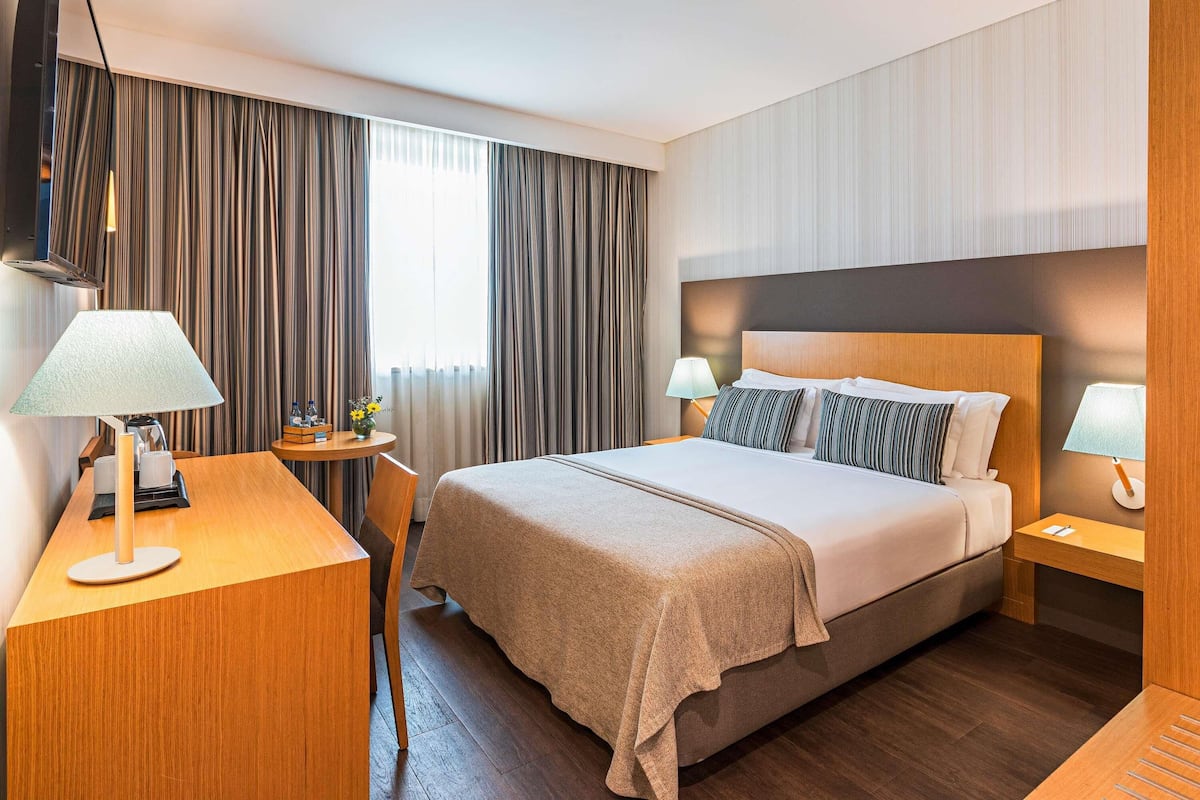 A hotel room with a bed, bedside tables, a desk, and a window with curtains.