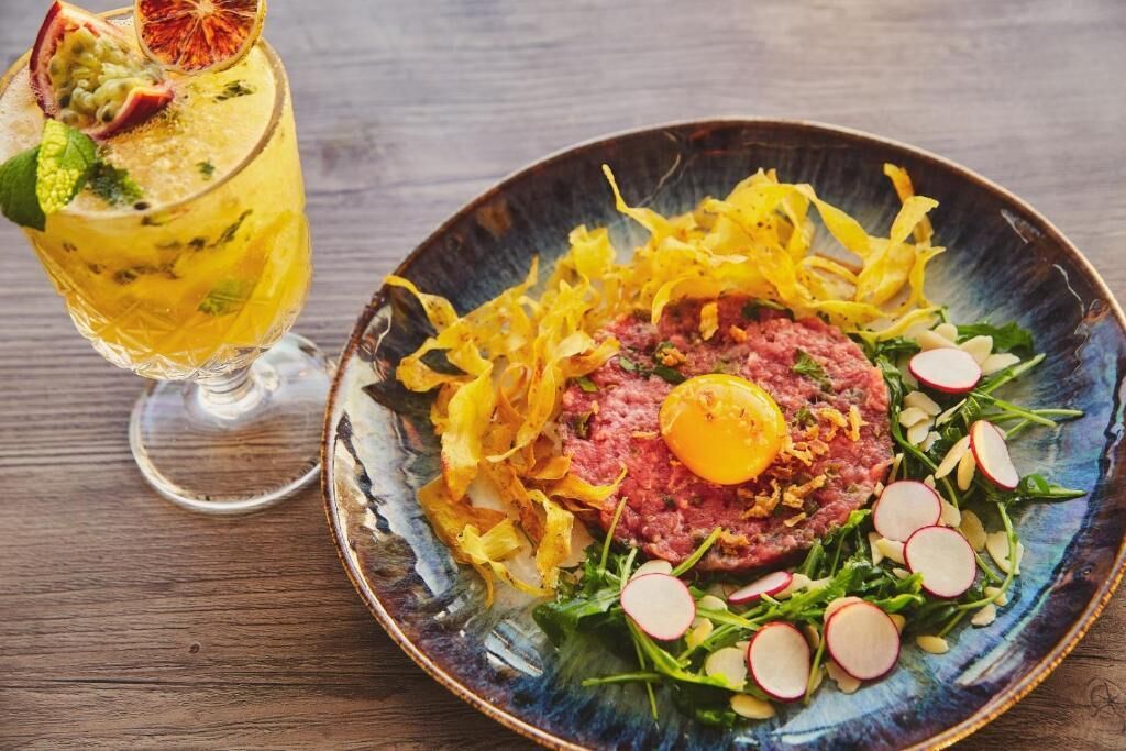 Ein Teller mit Steak-Tartare, verfeinert mit Ei, Radieschen und Grünzeug, sowie ein Cocktail mit Früchten und Minze.
