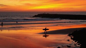 A beach scene at sunset with a person carrying a surfboard, waves, and a rocky shoreline. - Mercure Lisboa Almada (Almada)