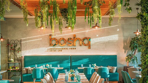 A restaurant interior with teal upholstered chairs, a long wooden table, hanging plants, and a wall with the name 'bosca'. - Mercure Lisboa Almada (Almada)