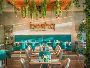 L’intérieur d’un restaurant présente des chaises recouvertes d’un tissu turquoise, une longue table en bois, des plantes suspendues et un mur sur lequel est inscrit le nom « bosca ».